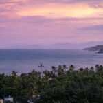 Beautiful Pink Sunset view over the ocean in Sayulita Mexico with mountain range and palm trees lining the coastline by Susan Paige Photography
